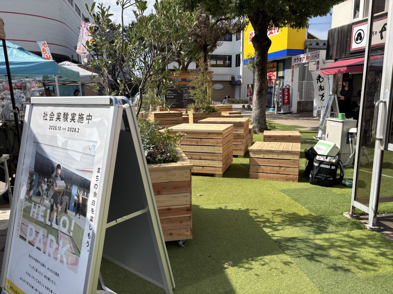 人工芝の上に並ぶ木製のテーブルやベンチ、プランター（加古川駅南広場）